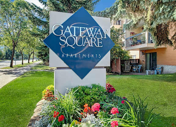 A welcoming community entrance sign here at Gateway Square surrounded by manicured landscaping, colorful flowers, mature trees, and nearby apartment buildings in a calm residential setting.