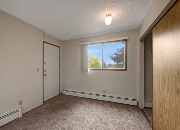 A bright, empty bedroom here at Gateway Square featuring soft neutral walls, plush carpet flooring, and a large window with vertical blinds that fills the space with natural light, plus a spacious closet and simple layout for a comfortable retreat.