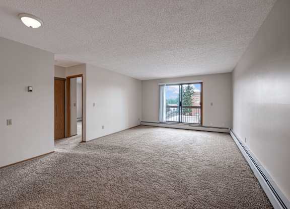 A bright living room here at Gateway Square featuring soft carpeting, crisp light walls, and a large sliding glass door that opens to a private balcony, filling the space with natural light and an airy feel.