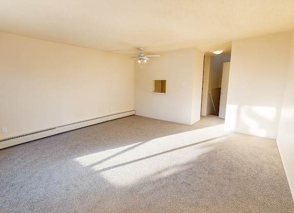 A sunlit living room at Lakewood Heights featuring soft carpet, neutral walls, baseboard heating, ceiling fan lighting, and an open, flexible layout with a pass-through detail that adds natural flow and everyday comfort.