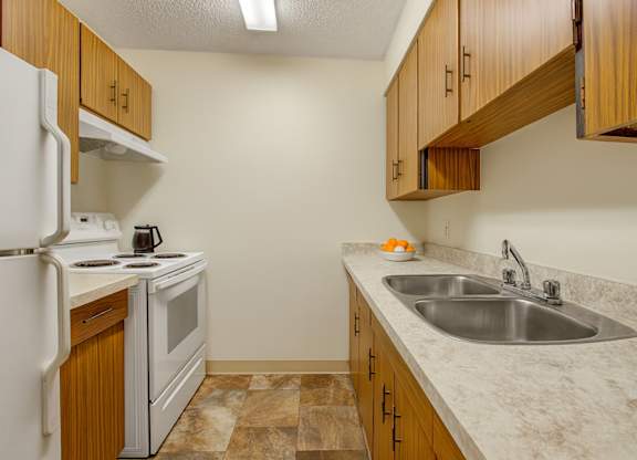 A clean, functional kitchen here at Lakewood Heights with warm wood cabinets, light countertops, a stainless steel double sink, white appliances, and efficient galley-style layout with bright overhead lighting.