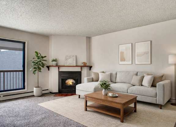 A warm, inviting living room here at Madison Park featuring a neutral sectional sofa with textured throw pillows, a wooden coffee table, soft area rug, and a cozy fireplace with a simple mantel, complemented by natural light from a sliding glass door leading to a private balcony.