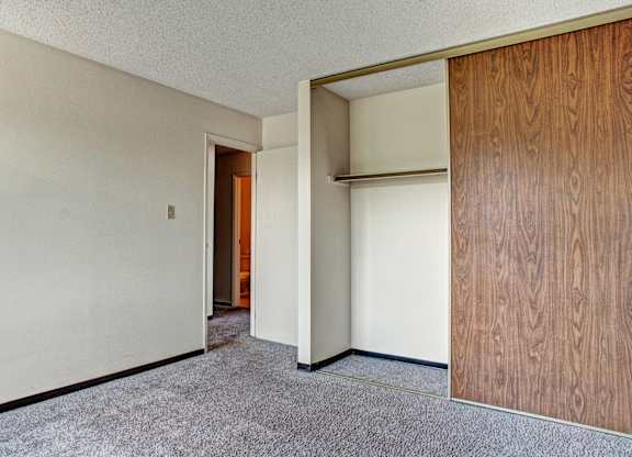 Empty room here at Madison Park Apartment Homes with beige walls, brown carpet, and an open white door leading to a hall. A closet with sliding wooden doors is on the right.