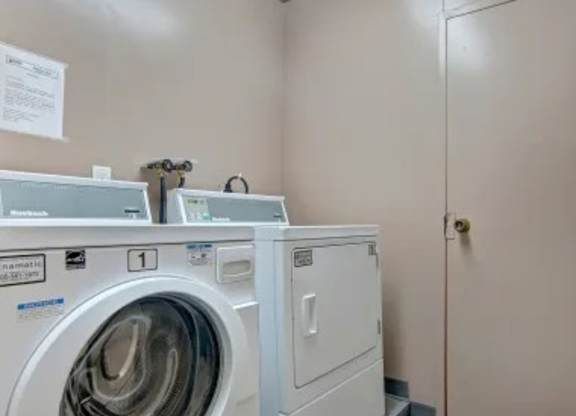 Laundry room here at Madison Park Apartment Homes with a white front-loading washing machine and dryer against a beige wall. Notice sign on the wall adds a utilitarian feel.