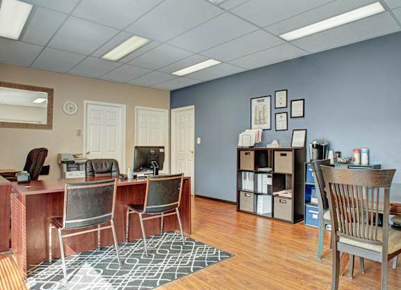 Leasing office here at Madison Park Apartment Homes with wood flooring and beige and blue walls. Contains desks, chairs, a computer, shelving with files, and framed certificates.