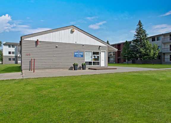 A well-maintained leasing office building here at Madison Park with neutral siding, a peaked roof, and a clearly marked rental center entrance. A concrete walkway leads to the door, with planters near the windows. Manicured green lawns, mature trees, and surrounding apartment buildings sit under a bright blue sky.