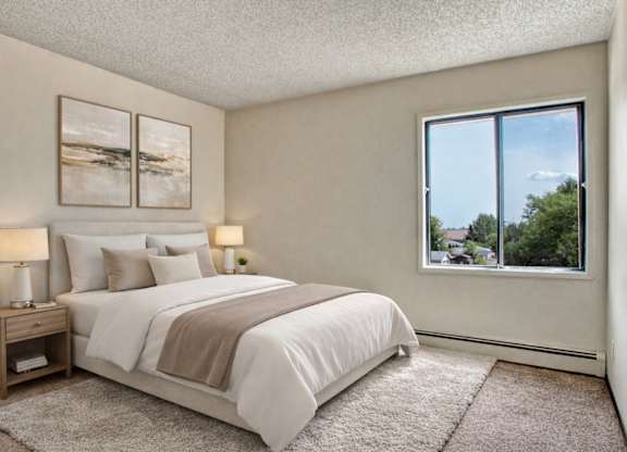 A bright, calming bedroom here at Madison Park featuring a neatly made bed with white bedding, layered neutral pillows, and a soft throw, paired with matching nightstands and warm table lamps, framed by minimalist wall art, plush carpet, and a large window offering natural light and neighborhood views.