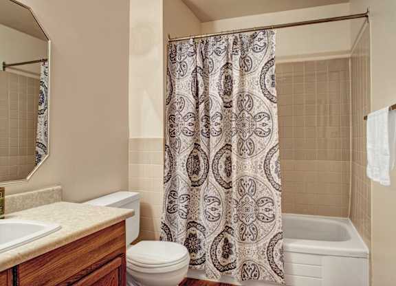 Bathroom here at Madison Park Apartment Homes featuring a tiled shower with a patterned curtain, a white toilet, a sink with a wooden cabinet, and a towel on a rack. Clean and tidy.