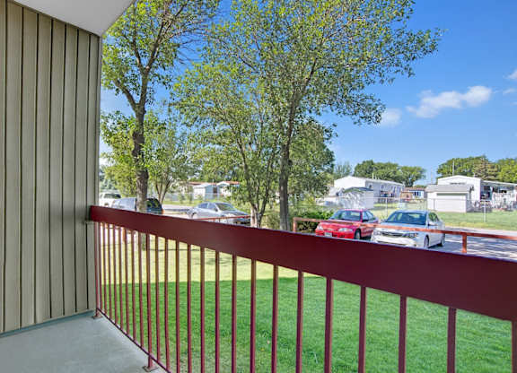 View from a balcony here at Madison Park Apartment Homes, with vertical siding and wooden railing, overlooking parked cars and trees. Mobile homes and blue sky complete the peaceful suburban scene.