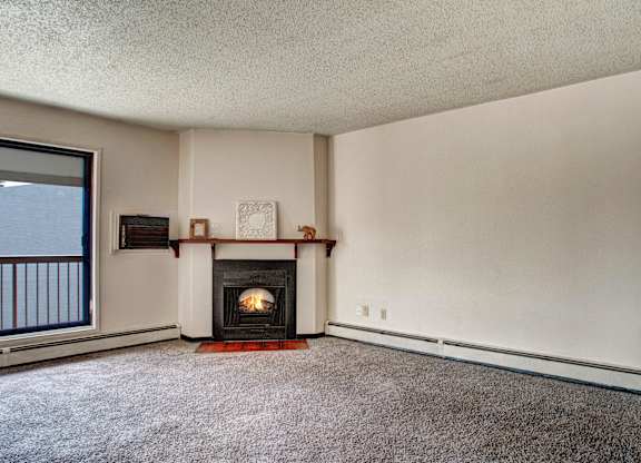 Spacious living room here at Madison Park Apartment Homes with beige walls and carpet, featuring a cozy fireplace with a wooden mantel. Large window provides natural light. Minimalist decor.
