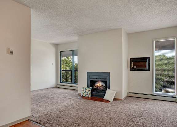 A cozy living room here at Madison Park Apartment Homes with a carpeted floor and a central fireplace framed by pillows. Large windows on each side let in natural light and offer views of greenery.