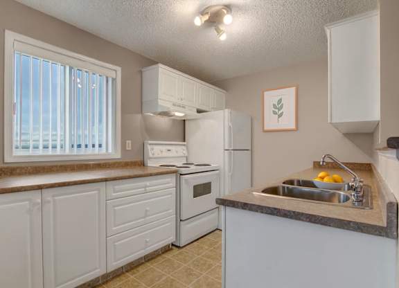 A bright kitchen here at Pointe at Applewood with white cabinets, brown countertops, and a double stainless steel sink. A window with vertical blinds brings in natural light, with a full appliance set and a clean, inviting layout.