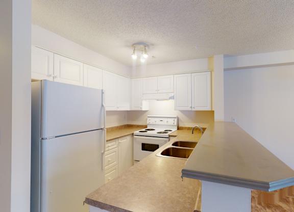 A functional kitchen here at Pointe at Applewood with white cabinets, brown countertops, and a double stainless steel sink. A breakfast bar overlooks the living area, with full-size appliances and a clean, open layout.