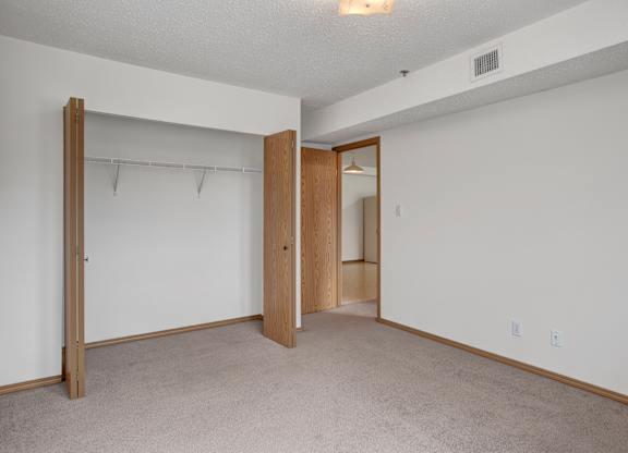 A spacious, empty bedroom here at Quail Ridge with soft carpet, clean white walls, and an open closet with built-in shelving for easy storage. A simple overhead light and warm wood trim add a bright, comfortable feel.