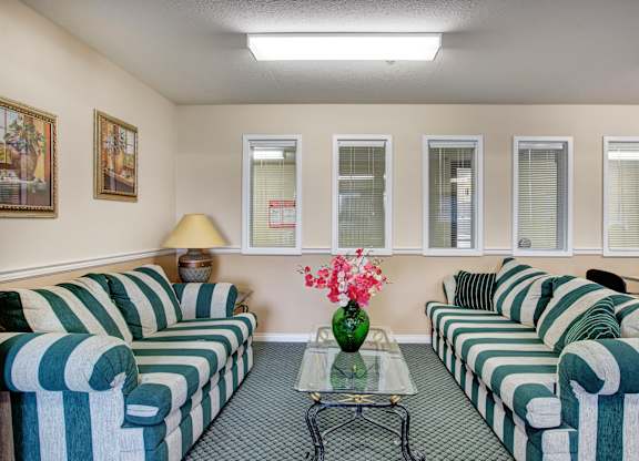 A cozy community lounge at **Quail Ridge** featuring two bold **green-and-white striped sofas** facing each other, a glass coffee table with floral decor, and soft overhead lighting. The space also includes framed wall art, a side table with a lamp, and interior windows that keep the room feeling open and connected.