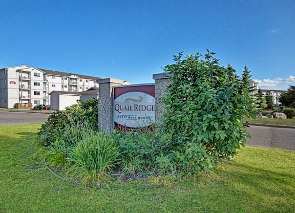 The Quail Ridge property entrance sign sits in a well-kept landscaped island with lush green shrubs and ornamental grasses, set against a bright blue sky with apartment buildings visible in the background.