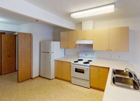 A bright kitchen here at Quail Ridge with light wood cabinets, white countertops, and a double stainless steel sink with generous prep space. A white refrigerator and electric range sit beneath ample cabinetry, and the room has a clean, open feel.
