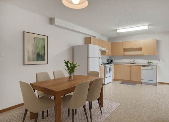 A bright dining area here at Quail Ridge with a wooden table, four upholstered chairs, and simple wall art for a clean, welcoming feel. A spacious kitchen sits just beyond with light wood cabinets, white appliances, and a neutral tile floor.