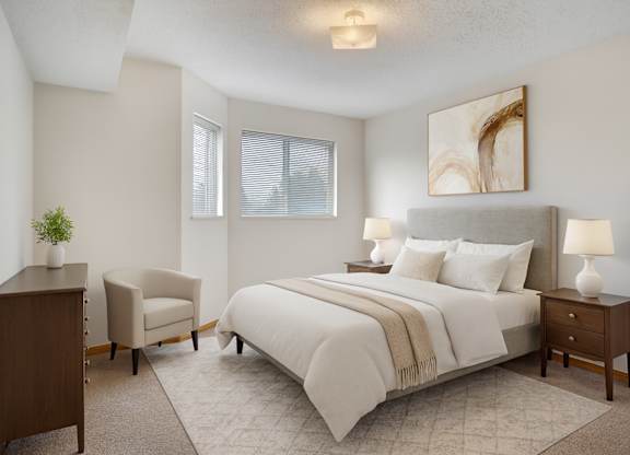 A bright bedroom here at Quail Ridge with soft carpet, neutral walls, and a cozy bed dressed in crisp white bedding and warm accents. A pair of nightstands with matching lamps and a cushioned chair create a calm, inviting space with gentle natural light from the windows.