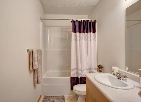 A clean bathroom here at Quail Ridge with a bright bathtub-shower combo, a modern striped shower curtain, and soft neutral walls. A simple vanity with a white countertop and sink sits beneath a large mirror, creating a fresh and functional space.