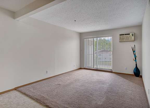 A spacious, empty living area here at Quail Ridge with soft beige carpet and clean white walls for a bright, open feel. A large sliding glass door with vertical blinds brings in natural light, and a built-in wall AC unit adds everyday comfort.