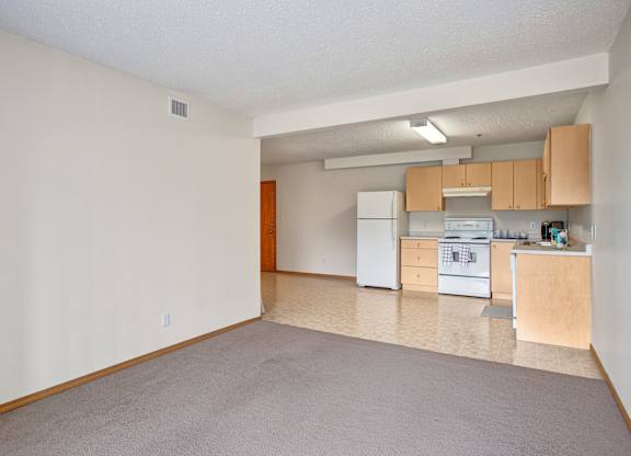 A bright, open-concept living area here at Quail Ridge with soft beige carpet flowing into a kitchen with light wood cabinets and neutral vinyl flooring. A white refrigerator and stove create a clean, classic look, and overhead lighting keeps the space airy and welcoming.