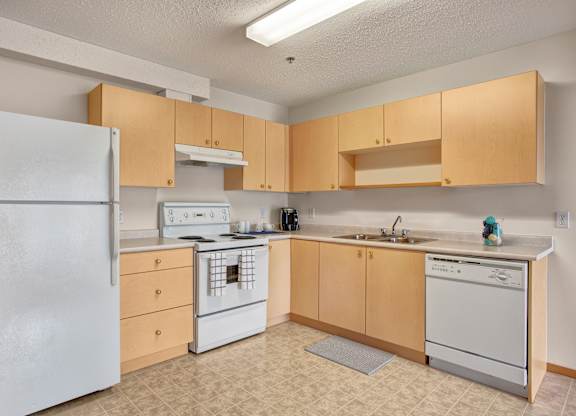 A bright, welcoming kitchen here at Quail Ridge with light wood cabinets, neutral countertops, and a classic white refrigerator and stove. A built-in dishwasher and stainless steel sink add everyday convenience, creating a clean and functional layout.