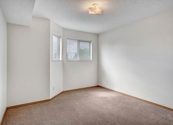 A bright, empty bedroom here at Quail Ridge with soft carpet, light walls, and natural light coming through wide windows with white blinds. A cozy corner nook adds extra space for a reading chair or small desk, creating a simple and inviting feel.
