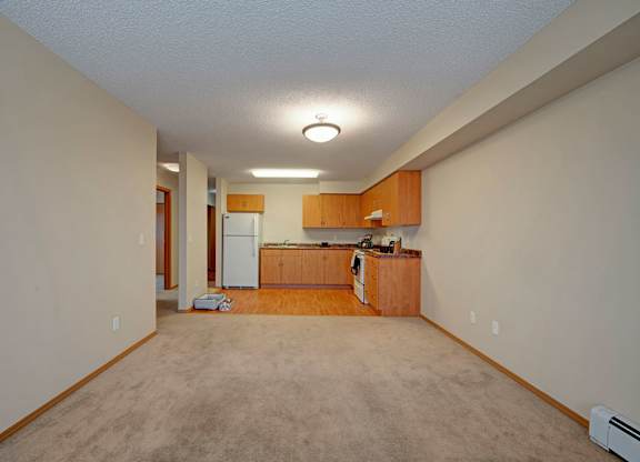 A spacious open-concept living area here at Sonora showcasing neutral carpeting, light-toned walls, and a seamless view into the kitchen with warm wood cabinetry, vinyl plank flooring, and overhead lighting, offering a versatile layout ideal for both relaxing and entertaining in a bright, welcoming apartment home.