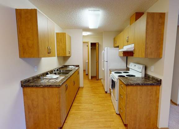 A bright galley-style kitchen here at Sonora featuring warm wood cabinetry, laminate countertops, white appliances, and wood-look flooring, with generous counter space, overhead lighting, and a functional layout that flows conveniently toward the adjacent living area for everyday cooking and dining.