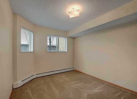 A cozy carpeted bedroom here at Sonora featuring soft neutral walls, plush flooring, baseboard heating, and two windows that allow natural light to brighten the space, with a simple ceiling light and a versatile layout suitable for a bed, dresser, and additional furnishings.