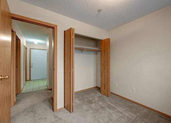 A cozy carpeted bedroom here at Sonora featuring neutral walls, an open wood-trimmed closet with double doors and hanging rod, and direct access to the hallway, highlighted by soft overhead lighting, warm wood accents, and a simple layout that allows for flexible furniture placement and everyday comfort.