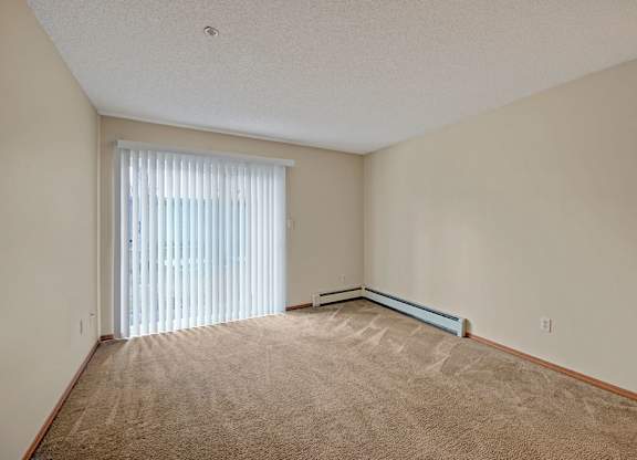 A bright carpeted living room here at Sonora featuring neutral walls, a large sliding glass door with vertical blinds that opens to a private balcony, baseboard heating along the wall, and an open rectangular layout that offers ample space for seating, media furniture, and everyday living.
