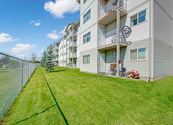 A landscaped exterior view here at Sonora showcasing a well-maintained green lawn alongside a multi-story apartment building with light-colored siding, private balconies on each level, patio seating with potted flowers, mature trees, and a fenced perimeter under a bright blue sky.