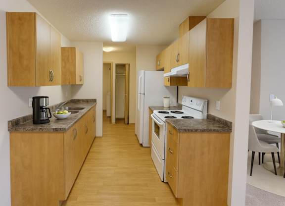 A warm, functional kitchen here at Sonora showcasing light wood cabinetry, laminate countertops, white appliances including a refrigerator and electric range, wood-style flooring, ample storage, and a galley-style layout that opens conveniently toward the adjacent dining area.