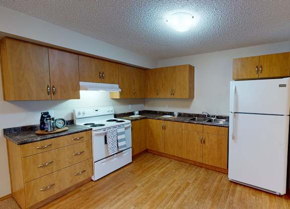 A bright, spacious kitchen here at Sonora featuring warm wood cabinetry, durable laminate countertops, white appliances including a full-size refrigerator and electric range, wood-style flooring, generous counter space, and an open layout that offers easy access to the adjoining hallway and living areas.