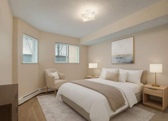 A cozy, light-filled bedroom here at Sonora featuring a queen-size bed with crisp white bedding, a soft neutral throw, matching bedside tables with warm lamps, plush carpet flooring, a comfortable accent chair, and windows with blinds that bring in natural light for a calm, inviting retreat.