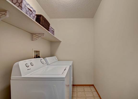 A convenient in-home laundry area here at Sonora featuring a full-size washer and dryer, built-in open shelving for storage baskets and supplies, neutral walls, and durable flooring that provides a practical and organized space for everyday laundry needs.