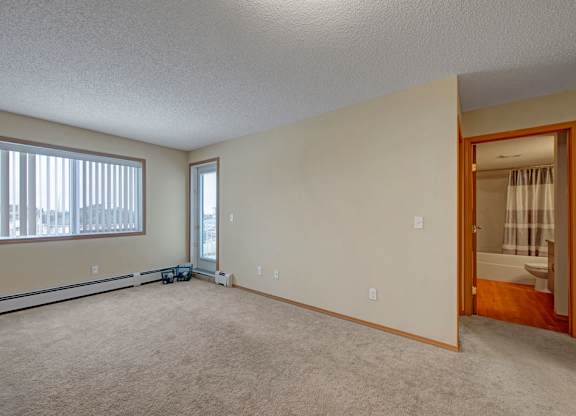 A spacious living room here at Sonora featuring soft neutral carpeting, light beige walls, and a large window with vertical blinds that brings in natural light, with a baseboard heater along the wall and an open doorway leading to a bathroom, creating a bright and versatile living space.