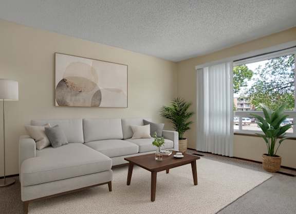 A bright modern living room here at Ventana featuring a light gray sectional sofa with neutral accent pillows, a wooden coffee table styled with minimal décor, a soft area rug, abstract wall art above the sofa, a tall floor lamp, and potted green plants near a large window with vertical blinds letting in natural light.