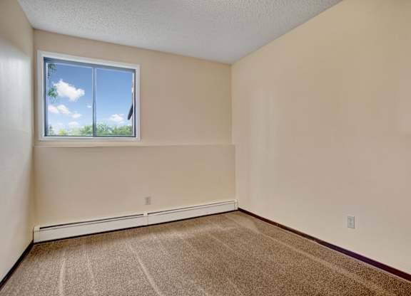 A cozy bedroom here at Ventana featuring soft neutral walls, plush carpet flooring, a baseboard heater along the wall, and a large window that brings in natural light and leafy outdoor views, creating a simple and comfortable space for rest.