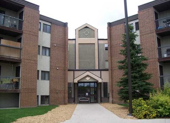 A welcoming apartment building exterior here at Ventana showcasing a multi-story brick and neutral-tone façade, a central covered entrance with glass doors, private balcony railings on each level, landscaped walkways, and mature greenery that create a well-maintained and inviting community setting.