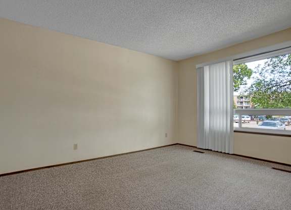 A spacious bedroom here at Ventana featuring neutral-toned walls, soft carpet flooring, a large window with vertical blinds that allow natural light, simple baseboard trim, and an open layout that offers flexibility for furniture placement and a bright, comfortable living environment.