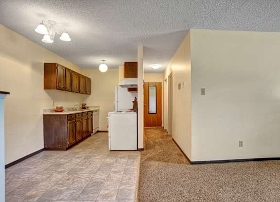 A bright open living space here at Ventana showcasing a combined living and kitchen layout with neutral walls, soft carpet and tile flooring, wood-style cabinetry, white appliances, overhead lighting, and a spacious hallway that creates a functional, comfortable flow throughout the apartment.
