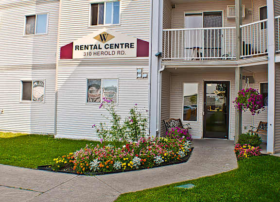 A welcoming rental office entrance here at Wellington Manor with a clearly marked Rental Centre sign and a covered walkway. A colorful landscaped flower bed and hanging baskets add vibrant curb appeal along the front path. A seating area near the door creates a friendly spot for guests and residents.