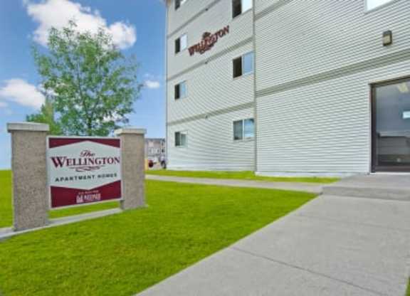 A bright exterior view here at Wellington Manor with a well-kept lawn and a clear community sign near the sidewalk. A multi-story apartment building with neutral siding creates a clean and welcoming look. A wide concrete walkway leads toward the entrance for easy access.