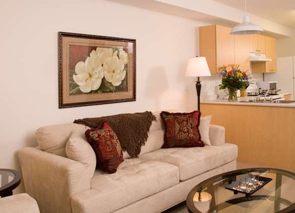 A warm and inviting living room here at Wellington Manor with a plush cream sofa, a round glass coffee table, and soft accent lighting. A floral wall art piece and patterned throw pillows add a cozy touch to the space. A bright open kitchen is visible in the background with light wood cabinets, a breakfast bar, and white appliances for a practical layout.