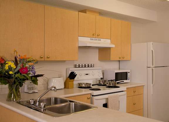 A bright kitchen here at Wellington Manor with light wood cabinets, clean white countertops, and a double stainless steel sink. A white stove with a vent hood, a microwave, and a full-size refrigerator provide a practical cooking setup. A wide breakfast bar counter adds extra prep space and a convenient spot for casual dining.