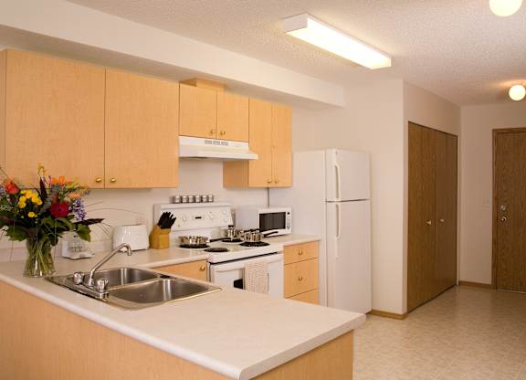 A spacious kitchen here at Wellington Manor with light wood cabinets, white countertops, and bright overhead lighting. A double stainless steel sink sits along a wide breakfast bar that offers extra counter space for cooking or casual dining. A full appliance set includes a white stove with vent hood, a microwave, and a full-size refrigerator for everyday convenience.