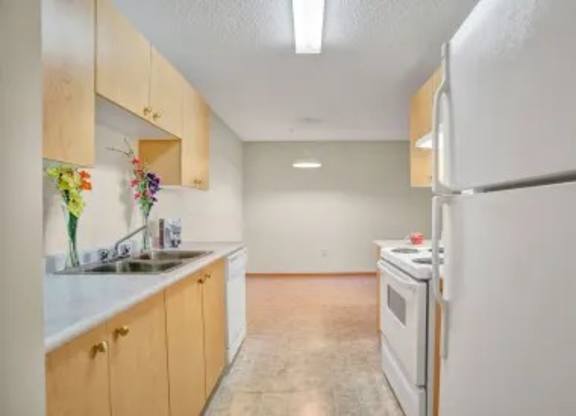A bright galley-style kitchen here at Wellington Manor with light wood cabinets, white countertops, and clean vinyl flooring. A full appliance set includes a white refrigerator, stove, and dishwasher for everyday convenience. A long counter and overhead lighting lead into an open dining area, creating a simple and functional layout.